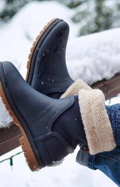 Navy waterproof boots with tan shearling lining and rugged soles resting on a snowy wooden railing outdoors.