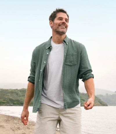 Person in green shirt over white T-shirt and khaki pants walking along a beach with hills in background.