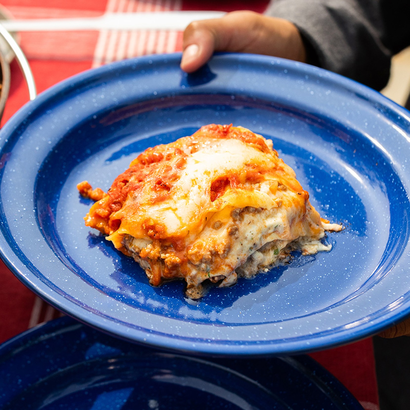 A hand holds a blue plate with a serving of lasagna topped with melted cheese and tomato sauce.