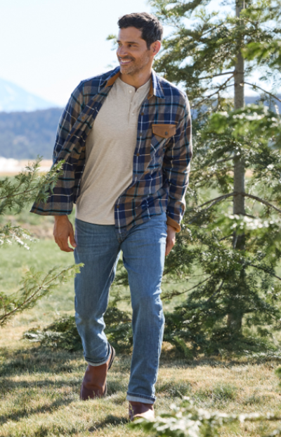 A person in a plaid shirt, beige top, and jeans walks outdoors among trees.
