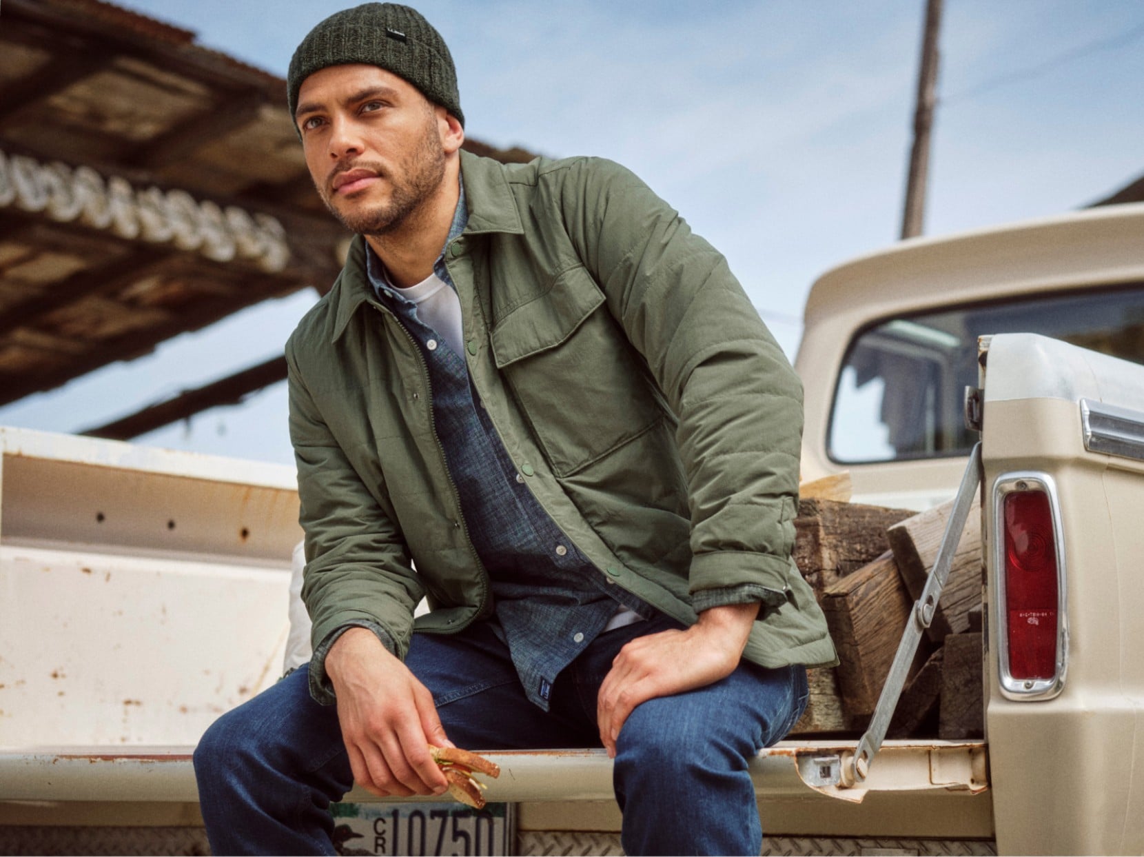 Man in a green jacket and knit cap seated on a pickup truck tailgate outdoors.