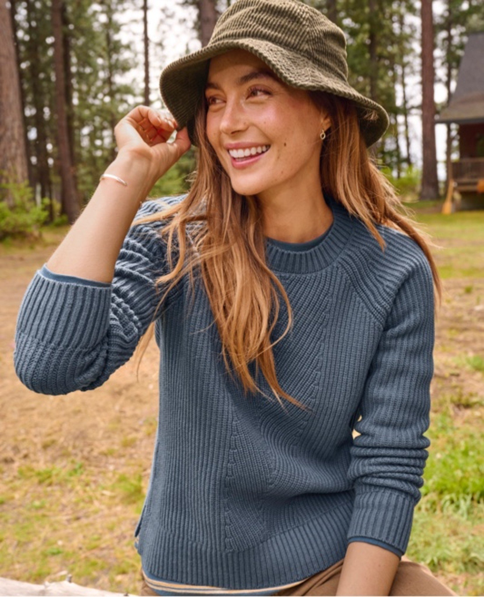 Person in blue sweater and green hat smiles outdoors near trees.