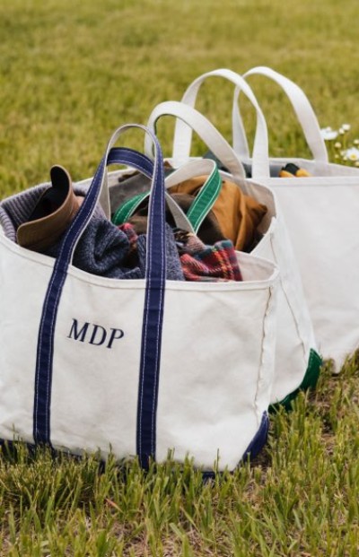 Two white canvas tote bags with colored handles sit on grass, filled with clothing; one bag is monogrammed M. D. P.