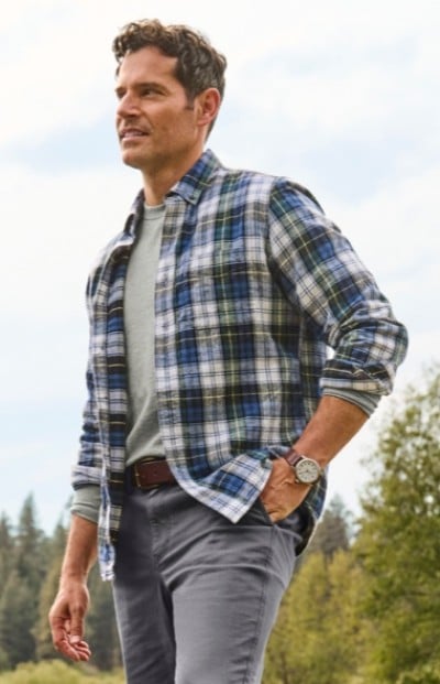 Man in blue and white plaid shirt over gray t-shirt stands outdoors with trees and sky in the background.