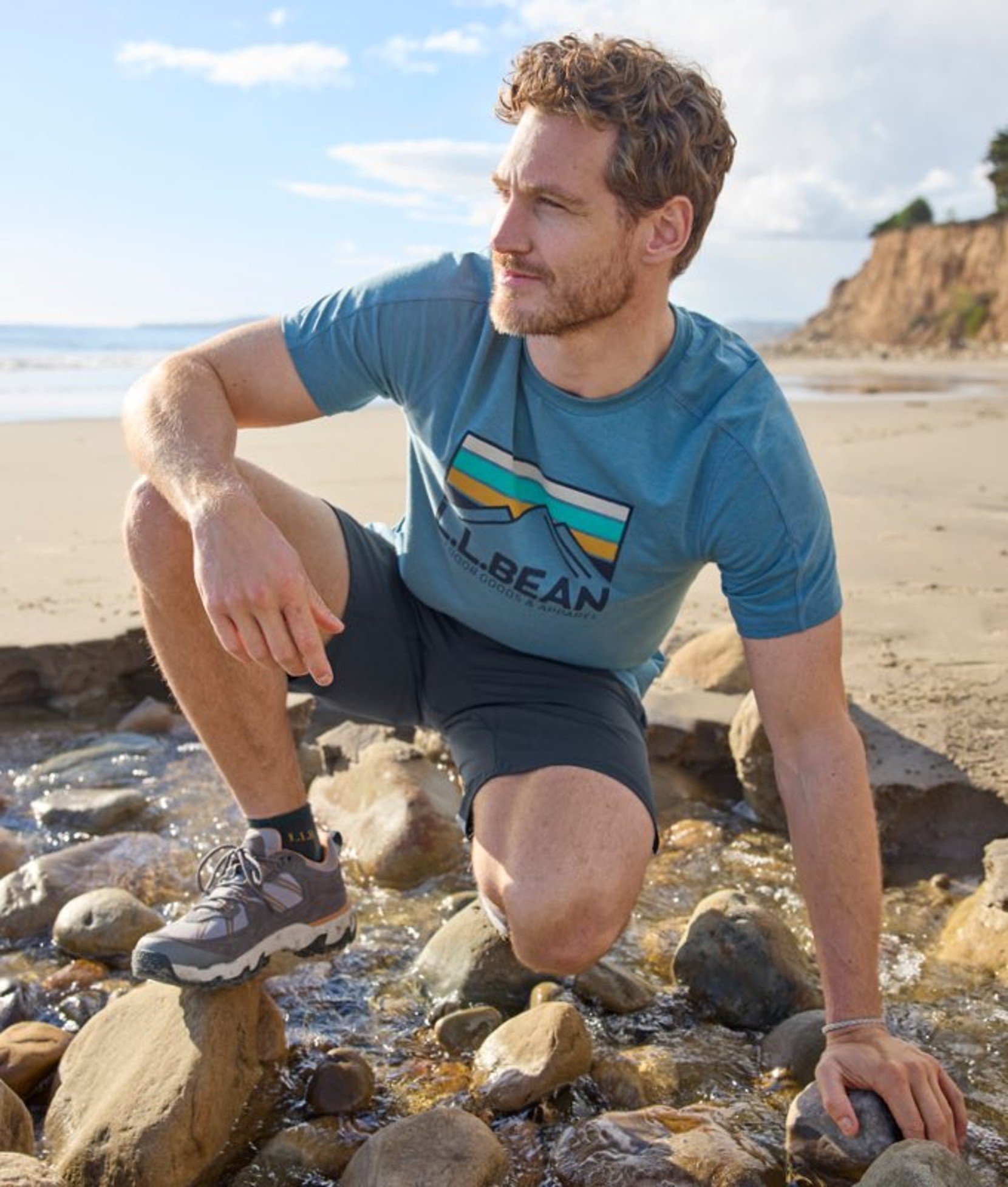 Person crouched on rocks wearing a graphic tee and shorts, with beach terrain and sky behind.