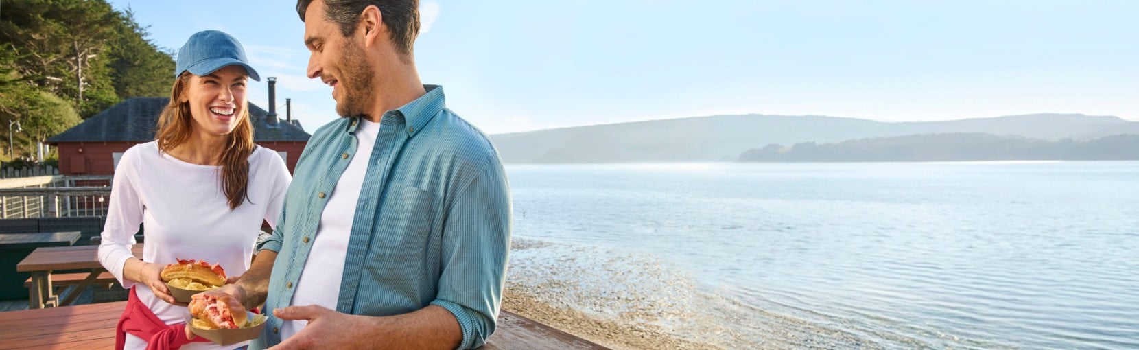 Two people holding lobster rolls near a waterfront with outdoor tables and distant hills in the background.