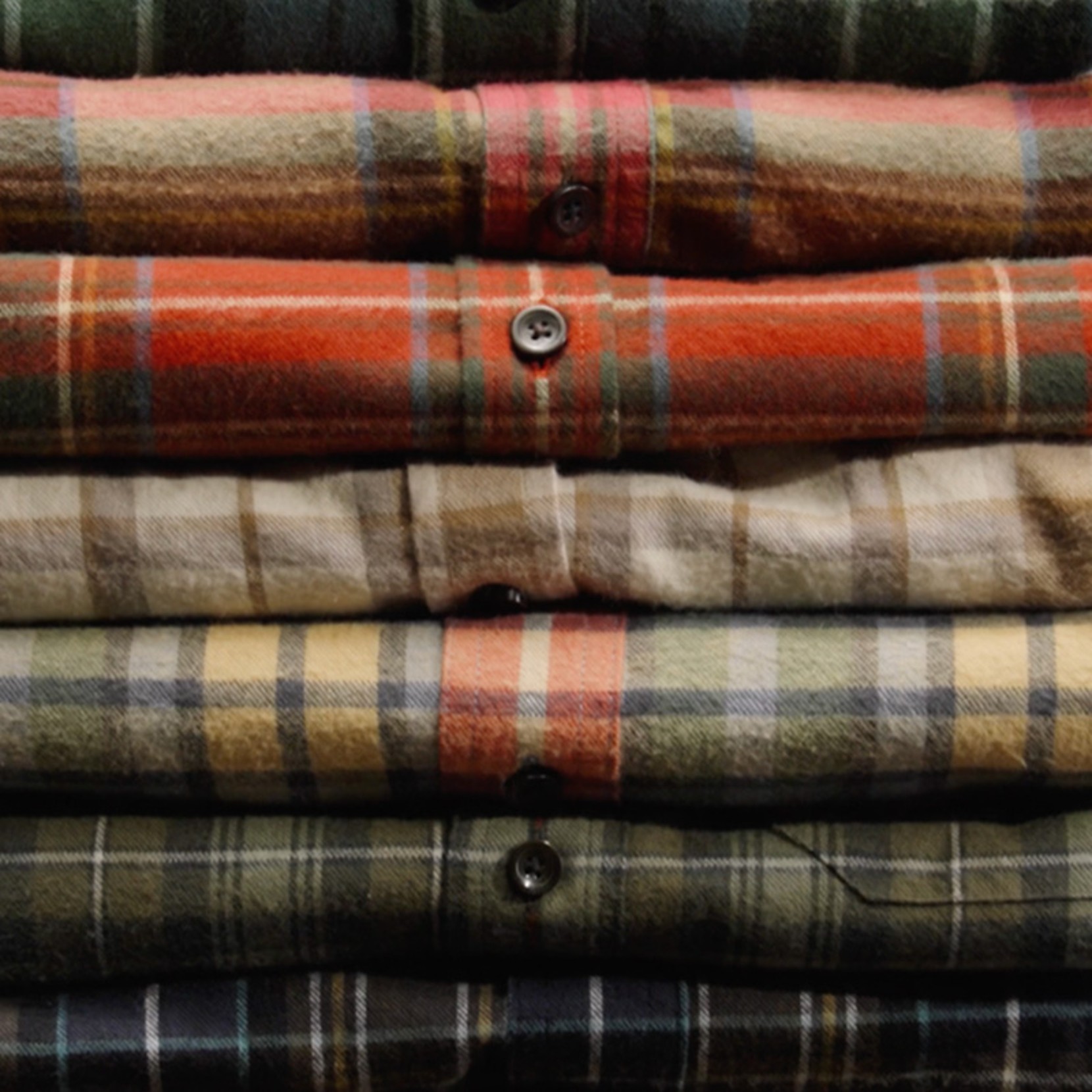 Stack of folded, flannel plaid shirts in red, green, and beige tones, neatly arranged with buttons visible. 