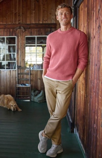 A person in a coral crewneck sweater and tan pants, stands in a rustic wooden cabin with a dog resting in the background.