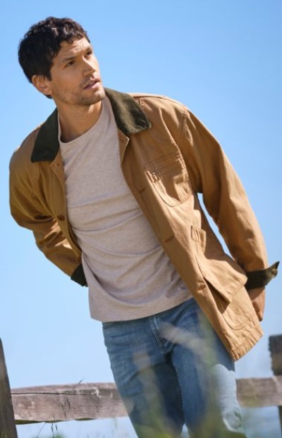 Person wearing a tan jacket over a white shirt walking outdoors on a sunny day.