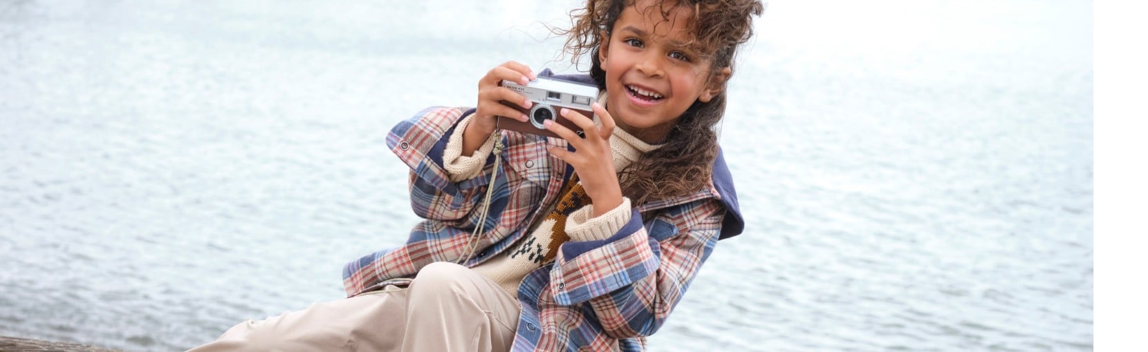 A child sitting outdoors near water, holding a small camera, wearing a plaid jacket and light-colored pants.