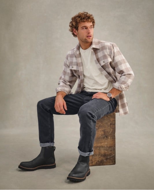 Person with curly hair in plaid shirt, jeans, and boots sits on wooden block, wearing a wristwatch.