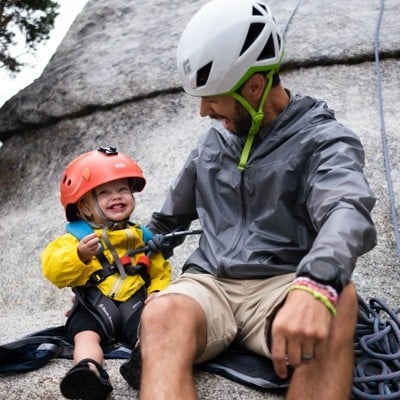 Man and young child in climbing gear sitting at the base of a rock wall.