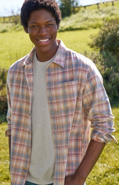 Person in plaid shirt and gray t-shirt stands outdoors on a sunny day with greenery in the background.