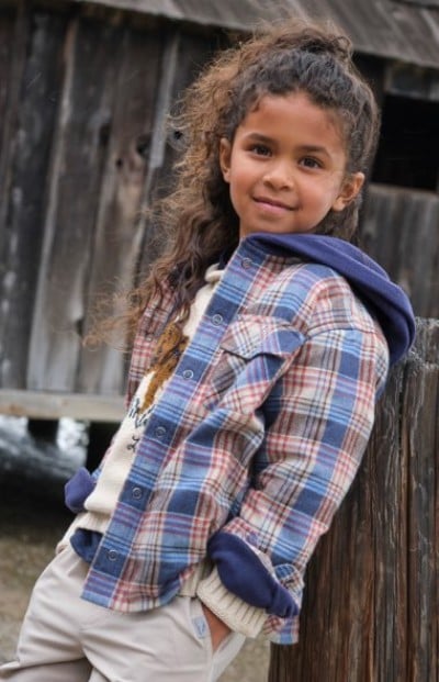 Child wearing a plaid hooded shirt leaning against a weathered wooden wall.