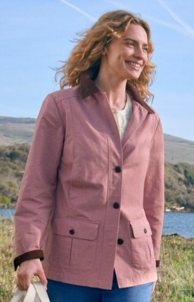Person wearing a pink button‑up jacket stands outdoors near grassy hills and water under a clear sky.