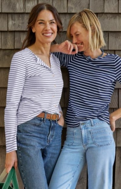Two people wearing striped long-sleeve tops and blue jeans, standing casually against a wooden shingle wall.