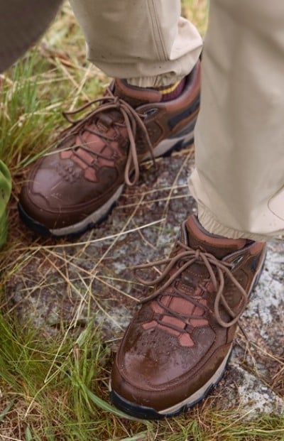 Brown hiking shoes with laces worn by someone in beige pants, standing on rocky ground surrounded by grass.