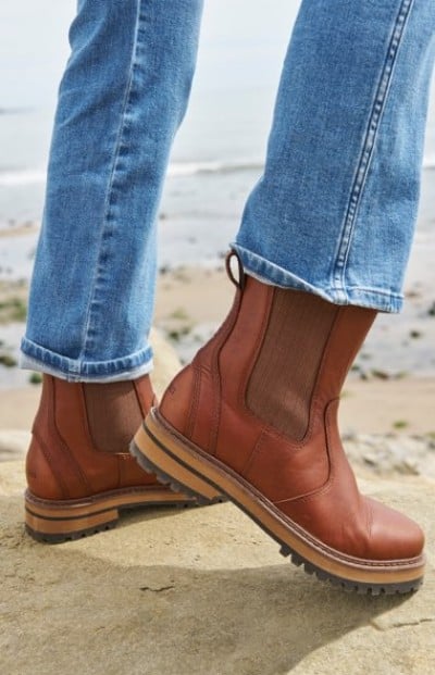 Brown leather Chelsea boots with chunky soles paired with blue jeans, standing on a rocky surface near the beach.