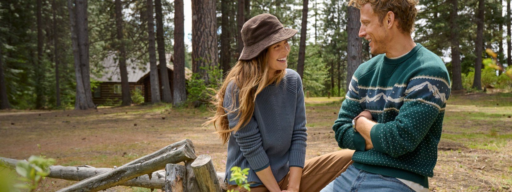 Two people in sweaters and hats sit on a wooden fence in a forest, with a cabin and tall trees in the background.