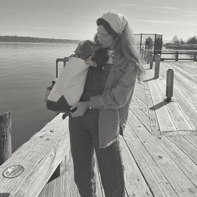 Woman holds a small dog wrapped in a blanket on a wooden pier by the water on a sunny day.