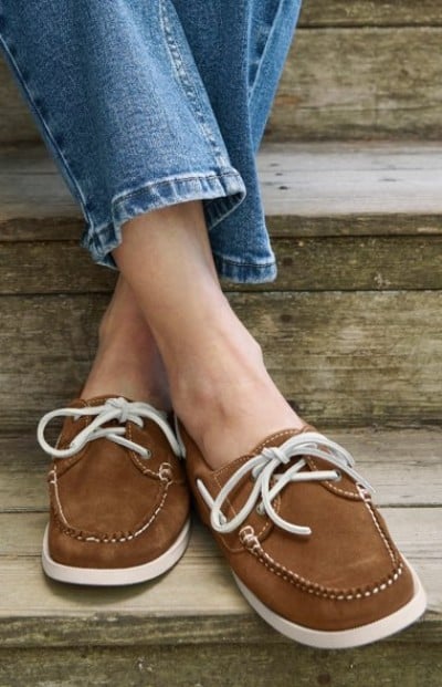 Close up image of a person in brown suede boat shoes with white laces resting on wooden steps, paired with blue jeans.