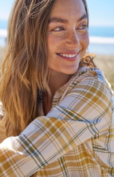 Woman with long hair in yellow plaid shirt smiles while looking to the side, with a blurred beach and ocean in the background.
