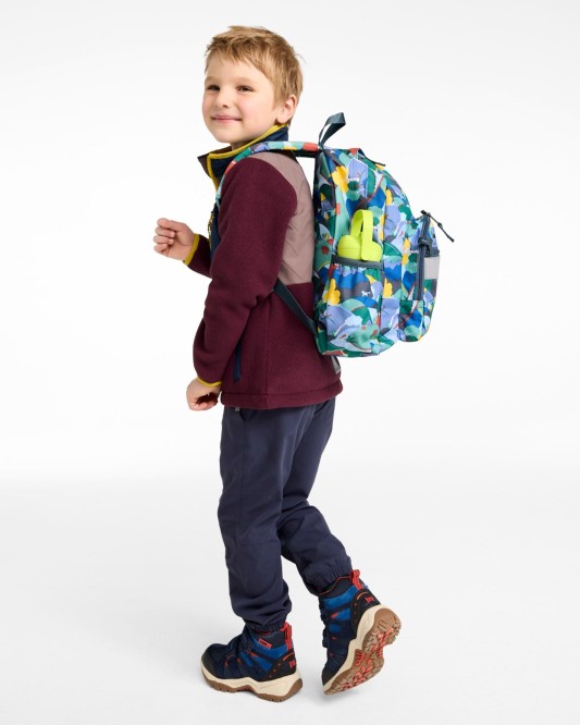 Young child in casual clothes walking and looking back, wearing a colorful backpack with side pocket items visible.