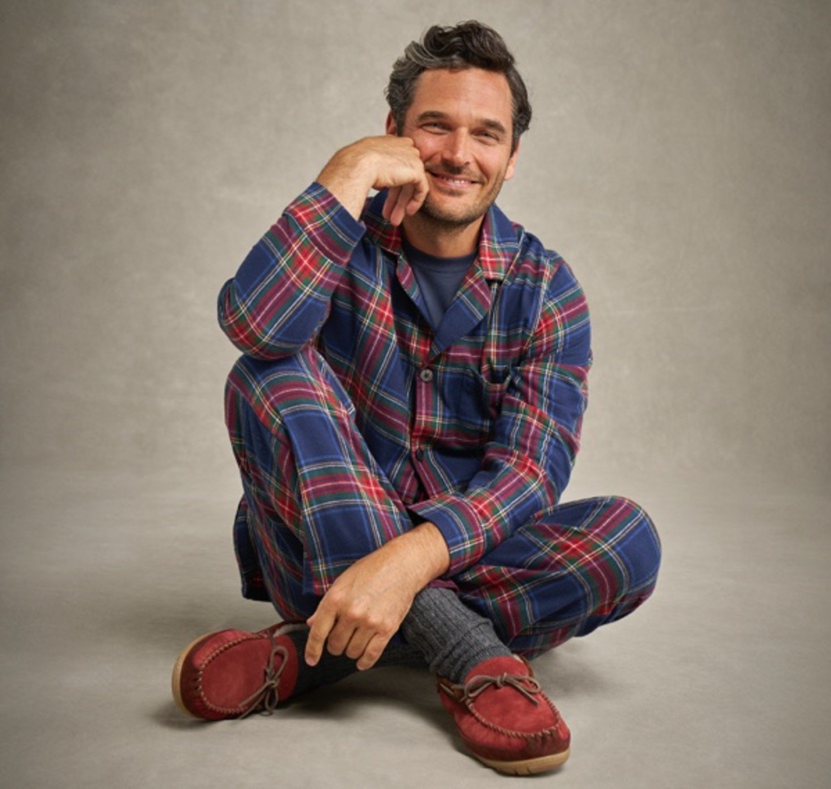 Person in plaid pajamas and red slippers sitting cross-legged on gray background.