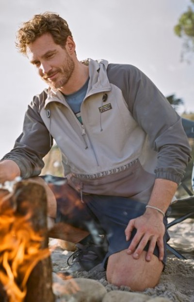 Person wearing a windbreaker sits tending a campfire on a beach at sunset.
