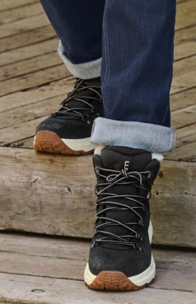 Black lace-up winter boots with white soles and brown tread, paired with cuffed dark jeans on a wooden deck.