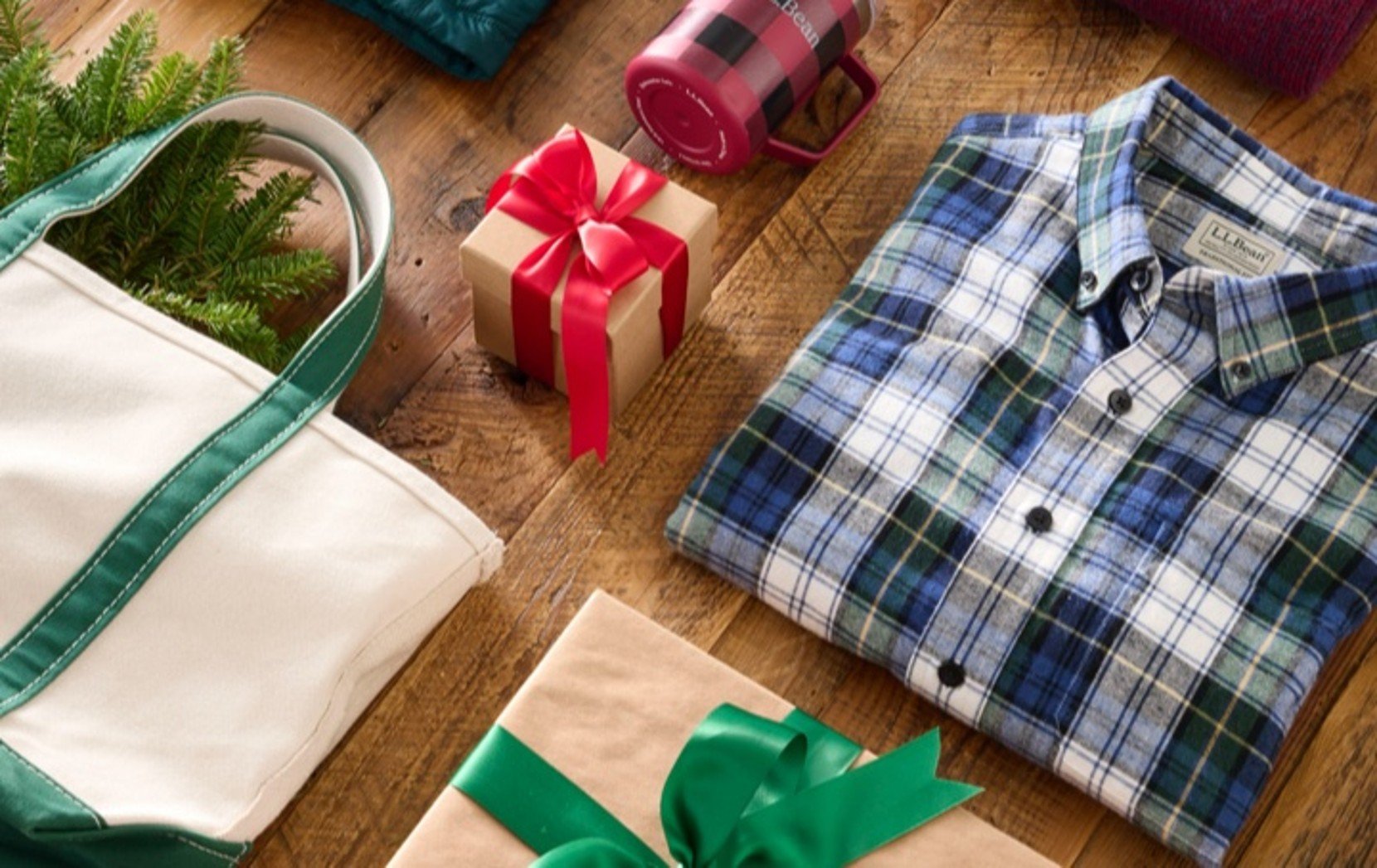 Winter clothes, wrapped gifts, a white tote bag, and pine branches arranged on a wooden floor in a festive holiday display.