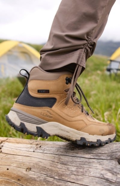 Person in brown hiking boots and pants steps on a log in a grassy outdoor area with tents visible in the background.