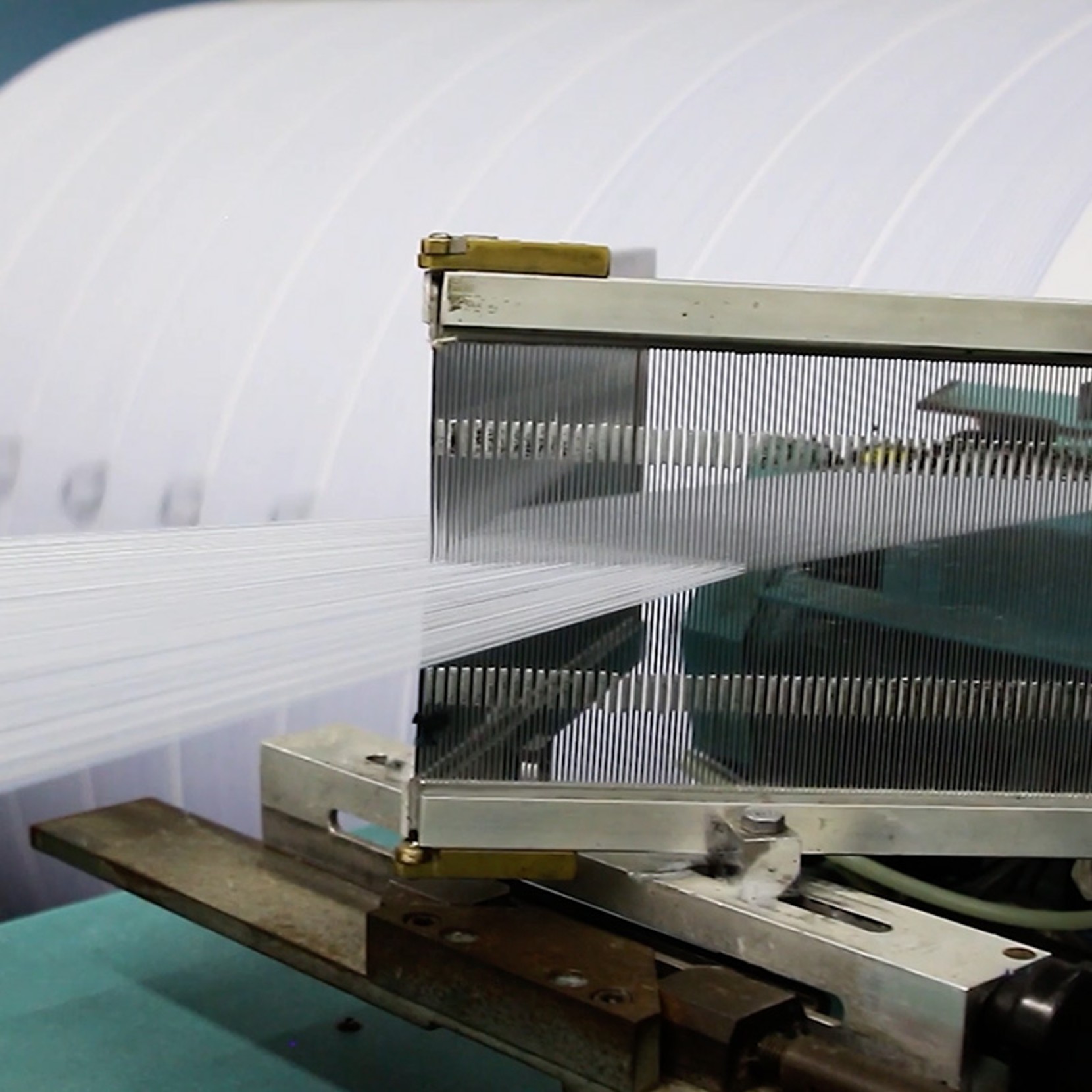 Weaving machine with white threads through metal combs, forming fabric; large roll of woven material in background.