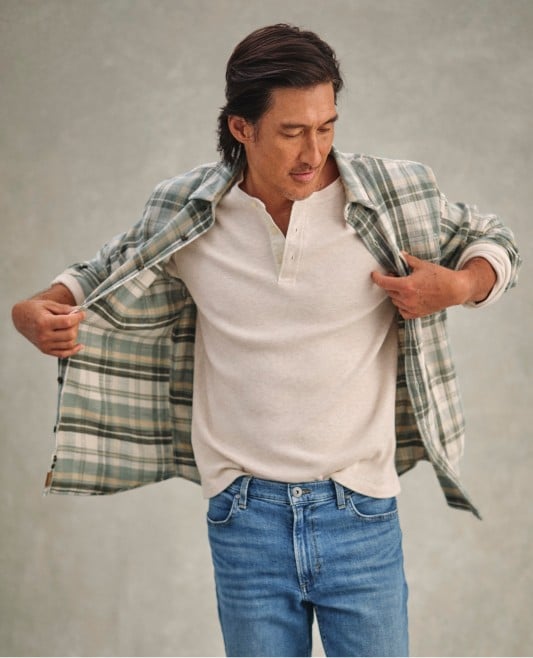 Person in green plaid jacket over beige henley and jeans looks down while adjusting jacket against gray background.