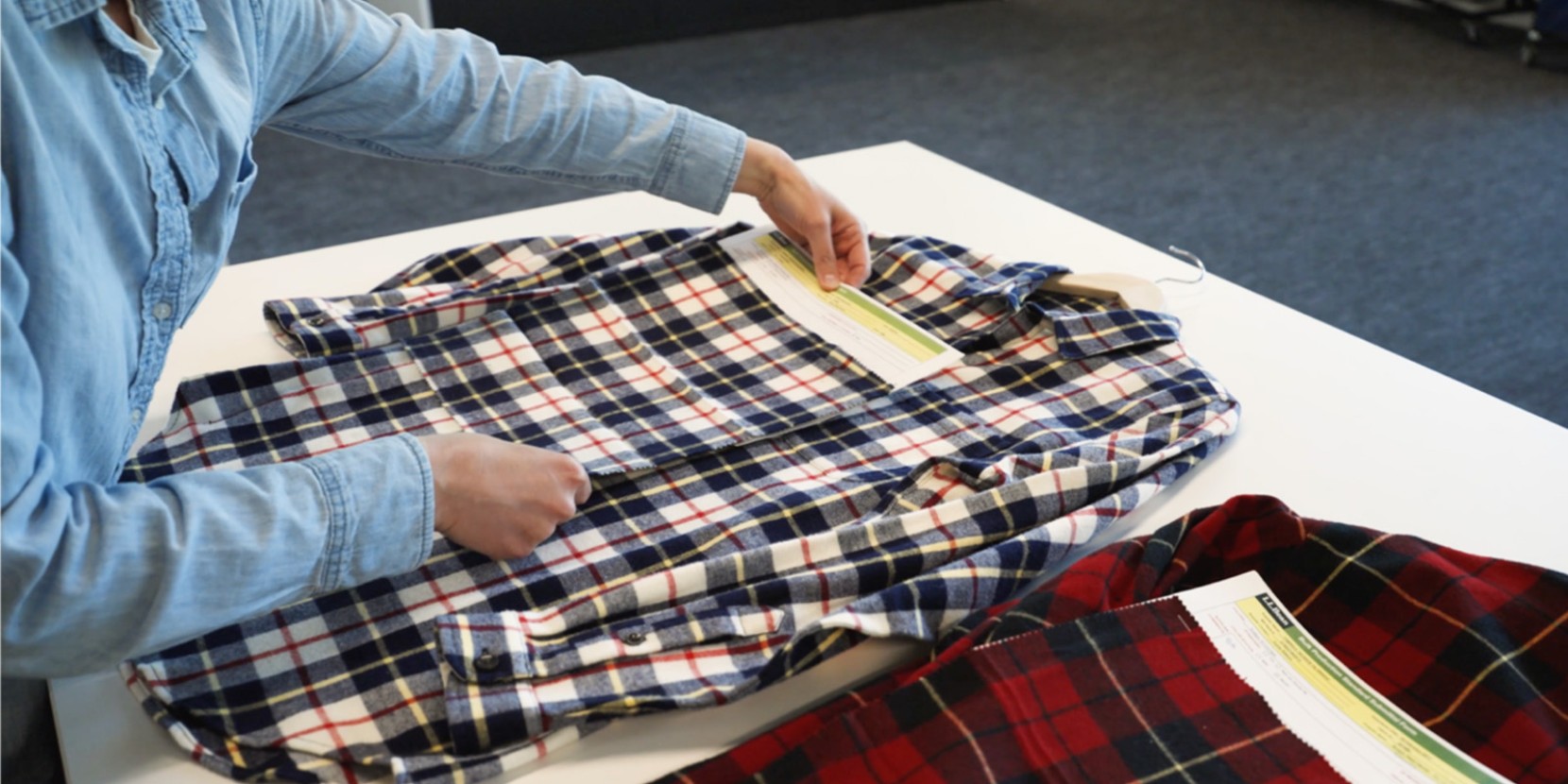 Person in a denim shirt folds a plaid shirt on a white table while holding an instruction sheet.