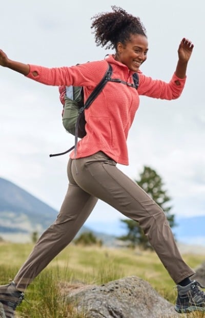 A woman in a pink top, tan pants and backpack hikes over rocks in a grassy area, with mountains and trees in the background.