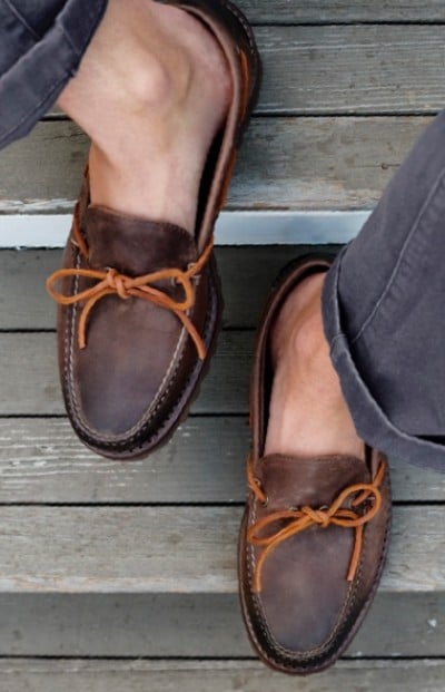Close up of brown leather moccasins with tan laces worn by someone in dark pants, standing on wooden steps.