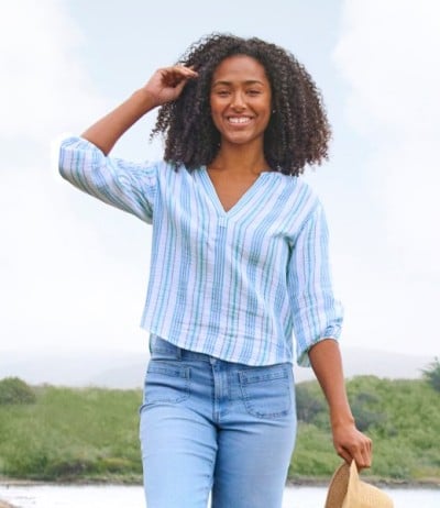 Person with curly hair wearing a blue striped blouse and jeans, holding a straw hat while walking near water and greenery. 