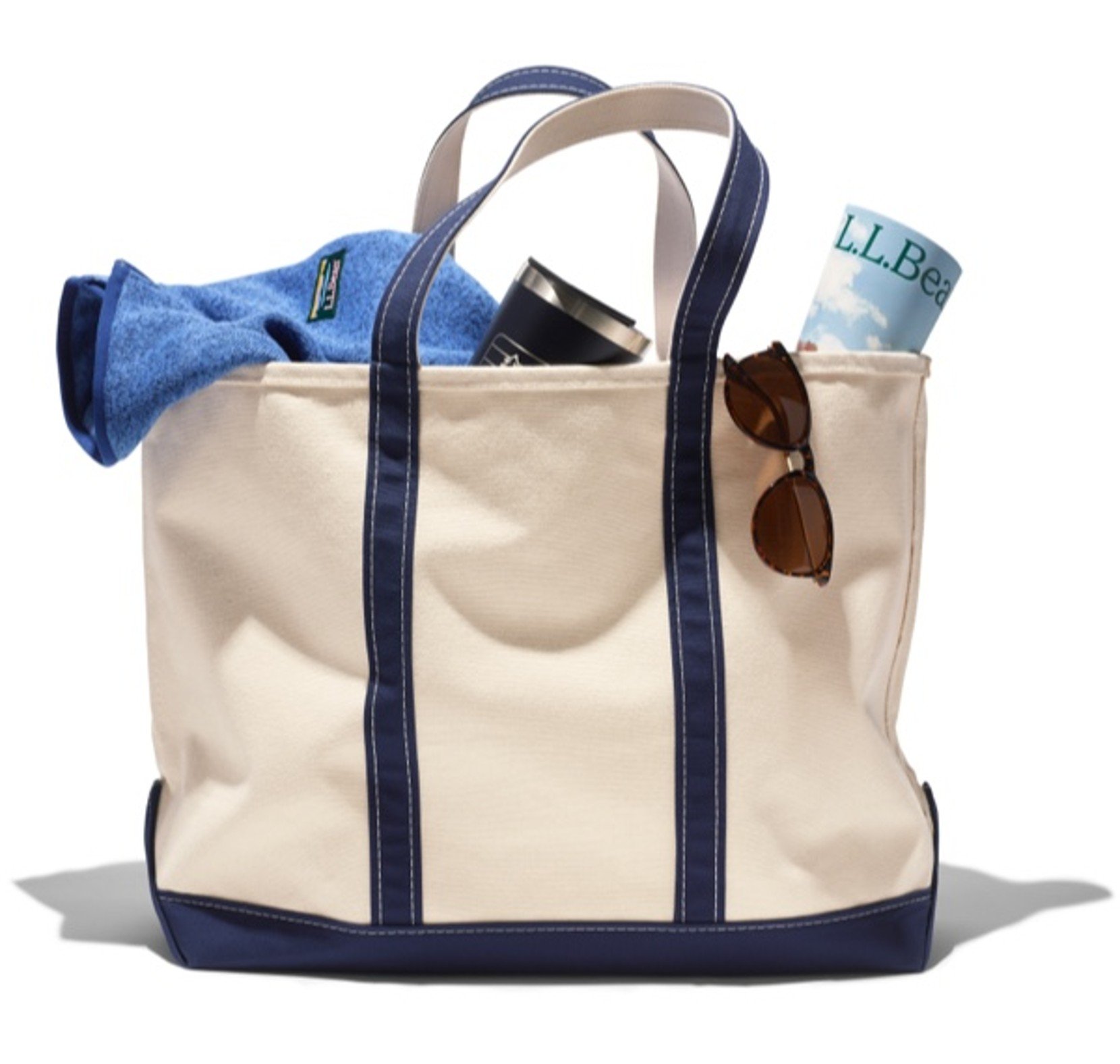 Canvas tote with blue trim filled with summer items like a towel, tumbler, and catalog.