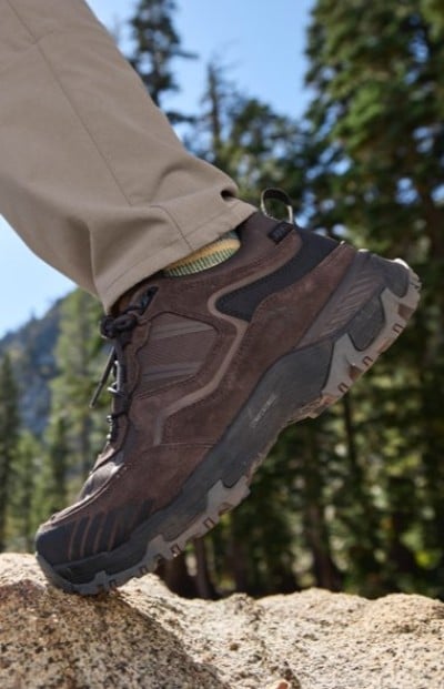 Brown hiking shoe with rugged sole and black accents, stepping on a rock in a forested mountain setting.