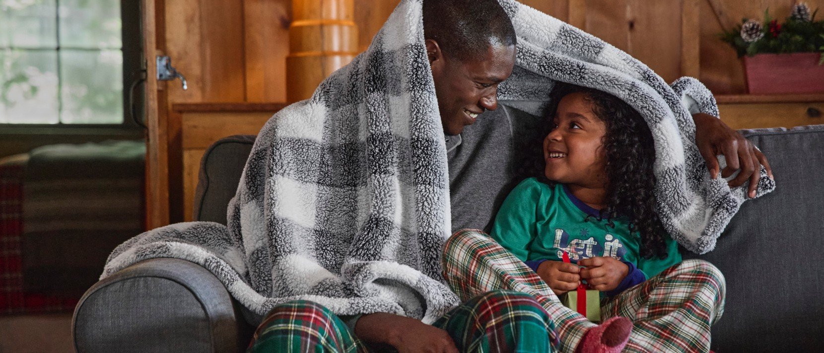 Two people in plaid pajamas sit on a couch, sharing a checkered blanket, with a lamp and wooden wall behind them.