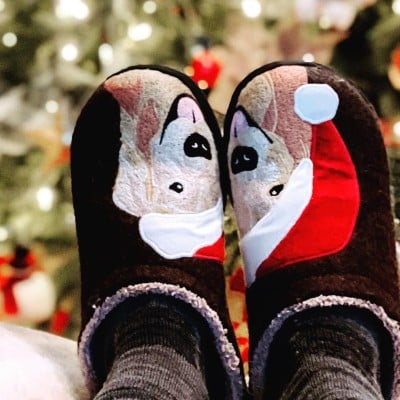 Feet wearing holiday motif slippers propped up on a chair arm, as seen from the person to whom the feet belong.