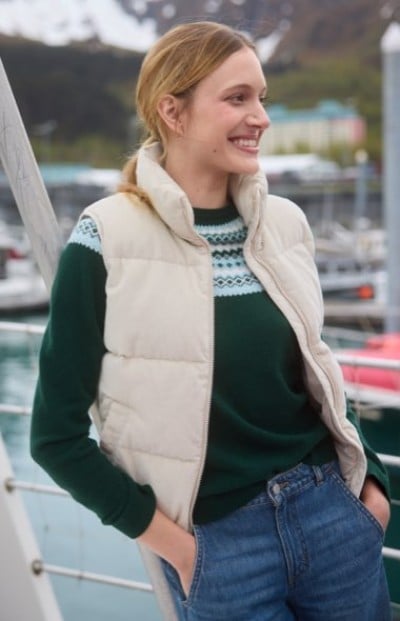 Person in a white puffer vest and green sweater standing near a marina.