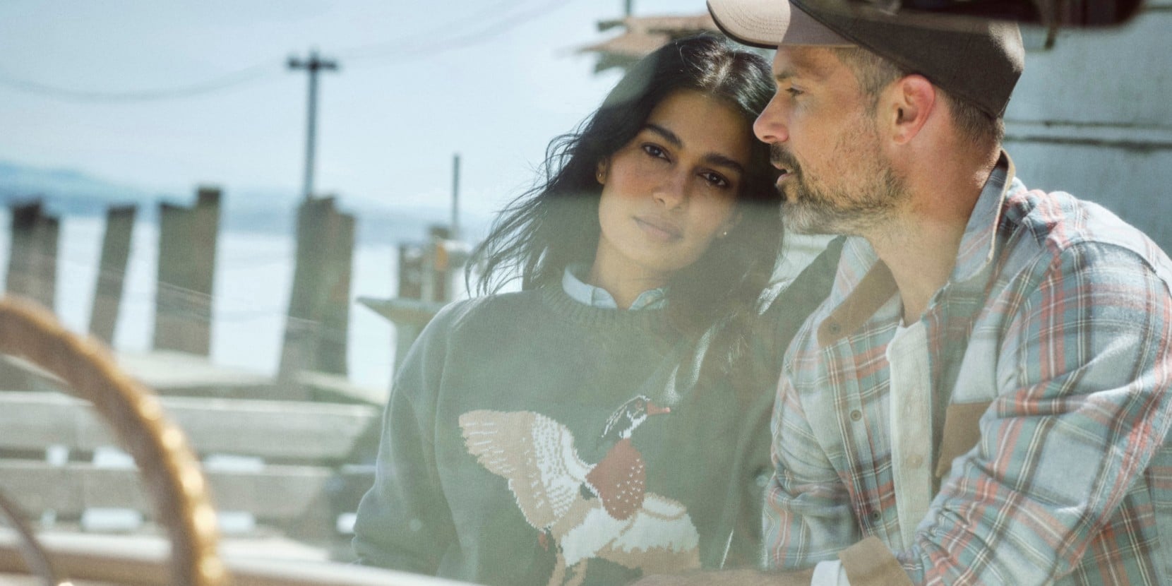 Person in bird-pattern sweater and person in plaid shirt sit together near a waterfront dock.