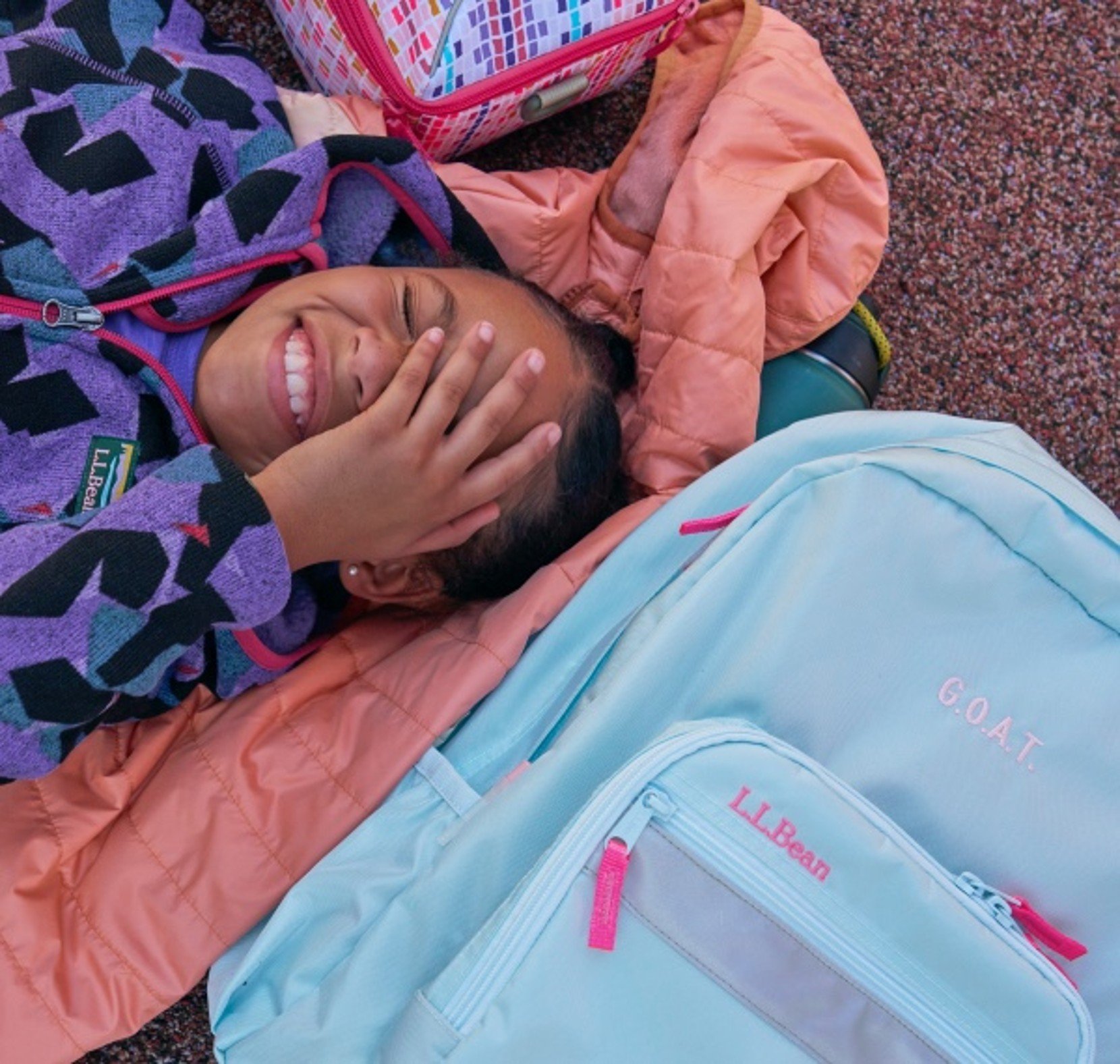 Child lying among colorful bookpacks and jackets, covering their face playfully.