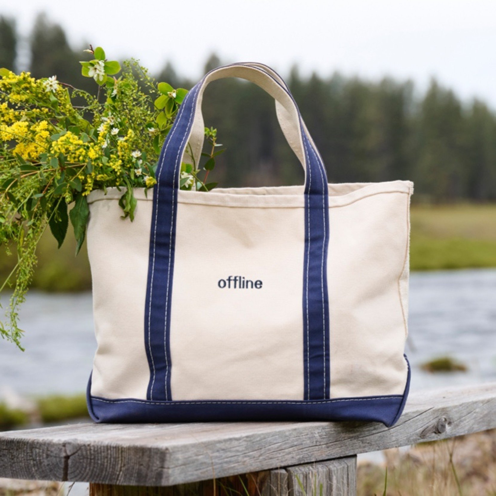 Beige tote with blue handles and 'offline' text sits on bench with yellow flowers, near river and forest.