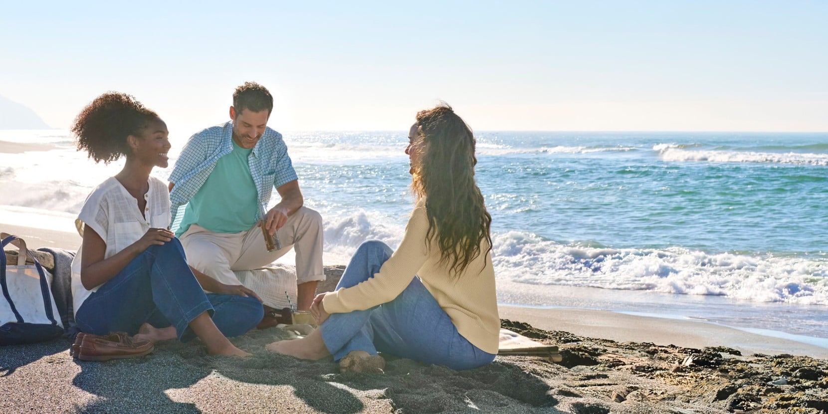 Three people sitting on rocks at the beach, wearing casual woven tops, with ocean waves and clear sky in the background.