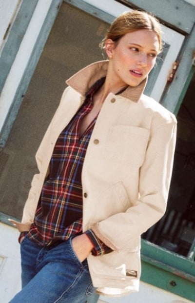 Person wearing a cream jacket over a plaid shirt stands beside a building entrance. 
