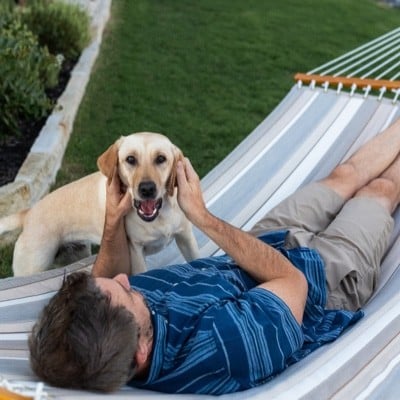 Man lying in a hammock petting a dog by his side.