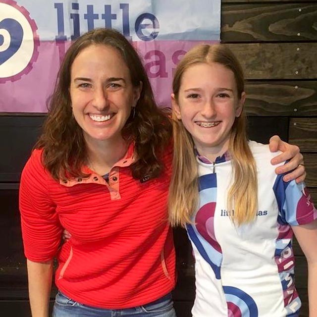 Lea and a young female cyclist in front of a Little Bellas banner.